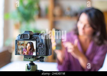 Une femme d'âge moyen qui diffuse en ligne depuis sa maison.Elle s'assoit avec un smartphone devant l'appareil photo et explique son sujet.Concept de technologie moderne. Banque D'Images