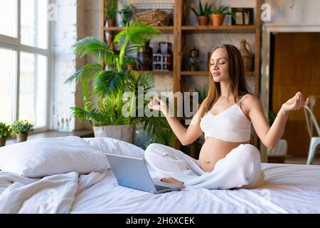 Une femme enceinte médite devant un écran d'ordinateur portable tout en étant assise sur le lit.Concept de santé mentale et de tranquillité pendant la grossesse. Banque D'Images