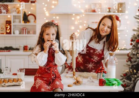 Une petite fille dans la cuisine du nouvel an est assise sur la table avec des biscuits dans ses mains, et sa mère prépare de la pâte. Banque D'Images