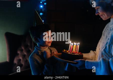 Joyeux enfant garçon avec les yeux fermés faire le souhait avant de souffler des bougies sur le gâteau de fête d'anniversaire dans la pièce sombre. Banque D'Images