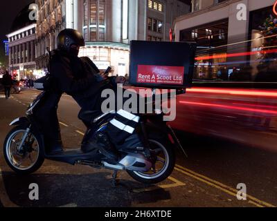 L'homme se penche sur son scooter en vérifiant son téléphone alors que les voitures passent en laissant des sentiers lumineux dans une longue exposition. Londres Banque D'Images