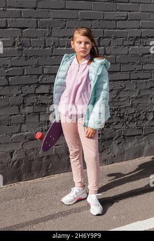 Petite fille dans des vêtements colorés se tient près de mur de brique grise avec un skateboard dans les mains Banque D'Images