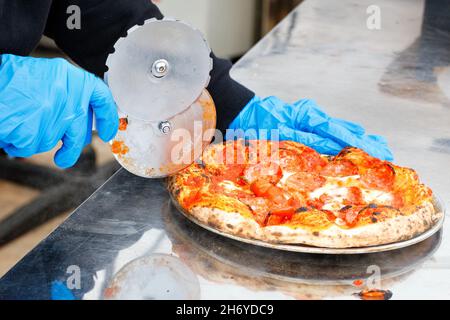 Un chef de rue en gants de latex bleu coupe une pizza cuite au four avec un couteau rond.Copier l'espace. Banque D'Images