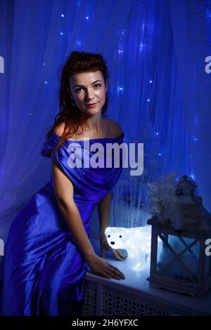 Jolie femme en robe bleue est assise par fenêtre sur le rebord de la fenêtre en soirée avec la lumière de nuit sous la forme d'un ours polaire.Une guirlande est suspendue à la fenêtre et brille d'une lumière douce.Ambiance festive et magique Banque D'Images