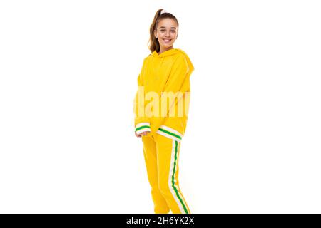 Photo de belle belle fille souriante mignonne avec des cheveux foncés avec queue de cheval dans élégant jaune tracksuit avec des rayures vertes isolées sur blanc Banque D'Images