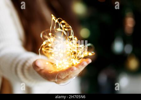 Miracle de Noël - une femme en chandail blanc tient la guirlande lumineuse de lumière LED dans les mains, arrière-plan bokeh. Banque D'Images