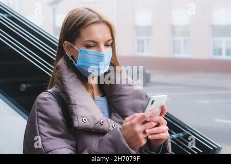 Jeune femme avec masque chirurgical protection du visage utiliser un smartphone tout en marchant à l'extérieur de la ville Banque D'Images