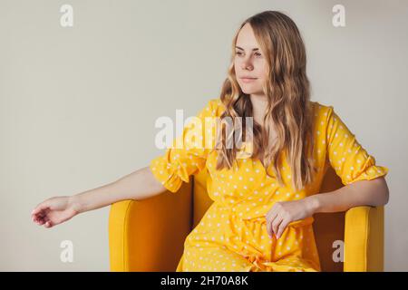 Portrait de belle jeune femme blonde souriante avec de longs cheveux bouclés dans une robe jaune rétro assis dans un fauteuil. Banque D'Images
