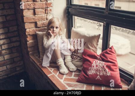 Une petite fille est assise sur un rebord de fenêtre en brique et soutient sa tête avec sa main entourée d'oreillers avec des inscriptions de Noël et fait des souhaits à San Banque D'Images