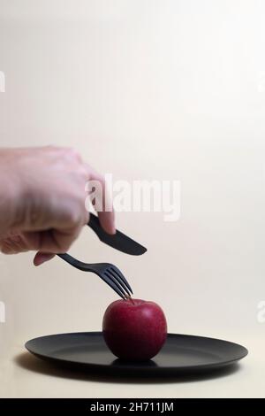 personne coupant la pomme rouge avec un couteau en plastique noir et une fourchette isolée sur fond clair.L'image contient de l'espace de copie Banque D'Images