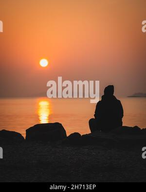 Coucher de soleil pittoresque au bord de la mer.Grand coucher de soleil sur l'horizon.Silhouette d'un homme assis seul sur un rocher en regardant le coucher de soleil orange.Lever du soleil le matin Banque D'Images