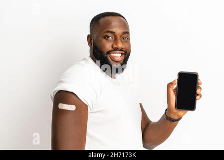 Homme afro-américain vacciné montrant un smartphone avec écran vierge, patch médical après injection sur l'épaule, homme noir recevant la dose de vaccin contre le covid, code qr, certificat de vaccin.Maquette Banque D'Images