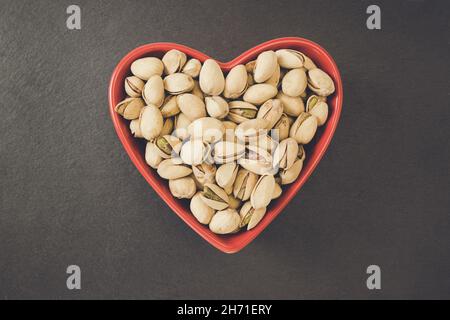 Pistaches salées rôties décortiquées dans un bol rouge en forme de coeur sur fond noir, concept de nourriture végétalienne saine et de collation.Gros plan, mise au point sélective, espace de copie Banque D'Images