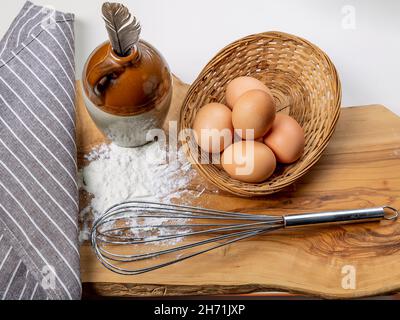 Un fouet de cuisine, de la farine, des œufs et une plume d'oiseau dans un récipient en céramique sur une planche à découper en bois Banque D'Images