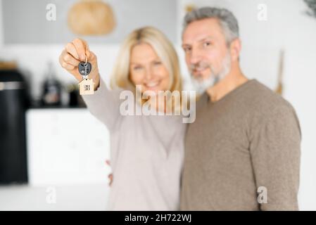 Attention sélective sur les clés avec porte-clés en forme de petite maison dans la main femelle, heureux joyeux couple d'âge moyen tient les clés de la nouvelle propriété, heureux acheteurs d'une propre propriété.Les conjoints obtiennent une hypothèque Banque D'Images