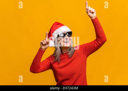 Femme mature en chapeau de père noël et lunettes de soleil dansant active isolée sur jaune, joyeuse femme moderne senior avec des cheveux gris appréciant la fête du nouvel an Banque D'Images