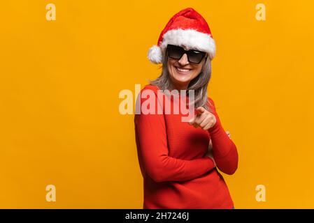 Une femme âgée enjouée dans un chapeau de père noël et des lunettes de soleil stylées pointe l'index vers l'appareil photo et rires, portrait une femme joyeuse d'âge moyen qui vous choisit.Concept de publicité de la nouvelle année Banque D'Images