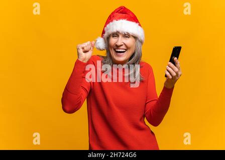 Surprise heureuse femme senior dans le chapeau de santa tenant un smartphone et se réjouit, la femme d'âge moyen tient un téléphone, a reçu un message inattendu ou une notification, regarde la caméra stupéfait, dit oui, isolé Banque D'Images