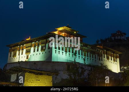 Le Rinpung ou Rinchen Pung Dzong illuminé la nuit à Paro, au Bhoutan.La tour de guet de Ta Dzong est au-dessus à droite. Banque D'Images