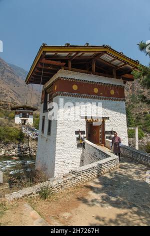 Homme bhoutanais dans un gho traditionnel devant un pont en chaîne de fer près du temple Tamchhog Lhakhang près de Paro, Bhoutan. Banque D'Images