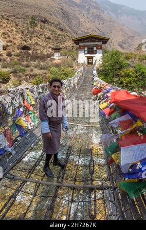 Un homme bhoutanais dans le gho traditionnel sur la chaîne de fer qui traverse la rivière Paro par le temple Tamchhog Lhakhang.Paro, Bhoutan. Banque D'Images