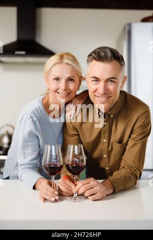Un couple d'âge moyen souriant se tenant ensemble dans la cuisine et buvant du vin rouge à la maison Banque D'Images