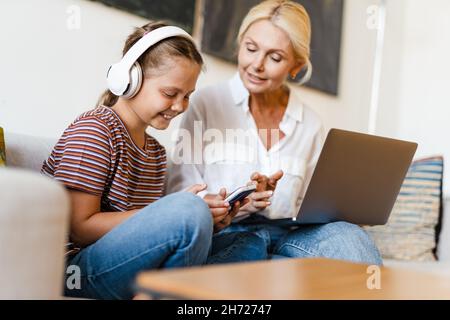 Mère et fille blanches utilisant un téléphone portable et un casque tout en étant assis sur un canapé à la maison Banque D'Images