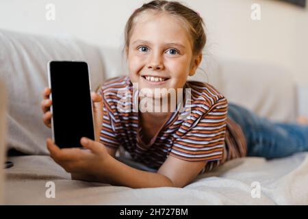 Jeune fille blanche souriant et montrant un téléphone portable tout en étant allongé sur un canapé à la maison Banque D'Images