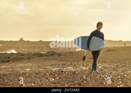 Jeune surfeur masculin avec surf marcher le long d'Avon Beach après avoir quitté l'eau au coucher du soleil en hiver, Royaume-Uni Banque D'Images