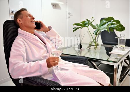Décontracté beau adulte 50 ans homme en éponge blanc peignoir assis sur un fauteuil avec un verre d'eau douce et parlant sur un téléphone portable au wel Banque D'Images