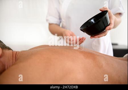 Gros plan d'un massothérapeute versant un peu d'huile du bol à l'arrière d'un homme mûr allongé sur une table de massage pendant une séance de massothérapie à h Banque D'Images