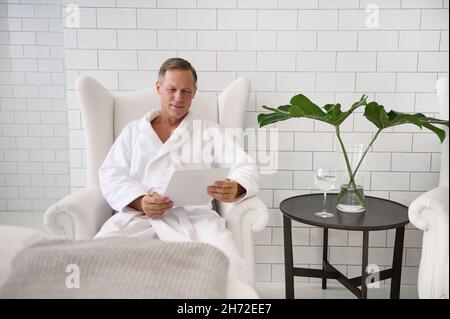 Beau homme d'affaires de 50 ans, avec succès, en tissu éponge blanc, portant un verre d'eau de citron et souriant, lisant le magazine pendant que r Banque D'Images