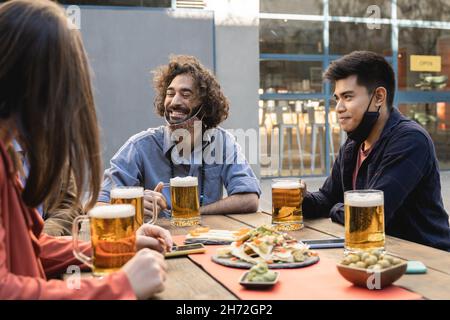 Les gens multiraciaux qui s'amusent à boire de la bière au bar de la brasserie en plein air tout en portant des masques de sécurité - Focus sur l'homme asiatique tenant le verre Banque D'Images