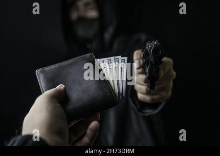 La main de l'homme tient le sac d'argent pour voler avec le pistolet.Vol à main armée.L'arme à feu est pointée à la caméra.Criminel dans le capot et le masque.Agression contre un homme non armé.Cent cinquante dollars dans un portefeuille ouvert. Banque D'Images