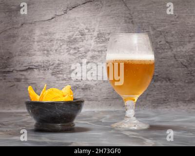 Un verre de bière et un bol en pierre avec des chips de pomme de terre sur fond gris. Banque D'Images