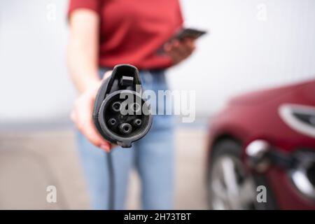 Une femme avec un smartphone montre la prise de charge électrique de la voiture à l'appareil photo Banque D'Images