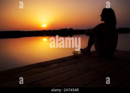 Silhouette d'une femme assise sur le quai en bois, se détendre avec une tasse de café et profiter d'un coucher de soleil doré sur l'eau. Banque D'Images