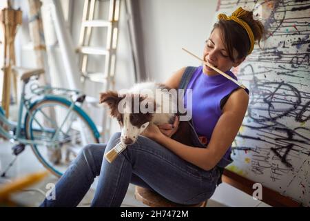 Une jeune femme artiste ayant un bon moment dans une atmosphère agréable dans son studio tout en jouant avec son chien.Art, peinture, amitié, studio Banque D'Images