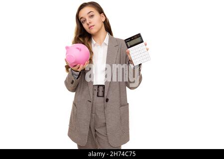 Photo de belle jolie petite fille brune avec de longs cheveux dans une veste à carreaux élégante, un pantalon à carreaux et une chemise blanche contient une calculatrice et un cochon rose Banque D'Images