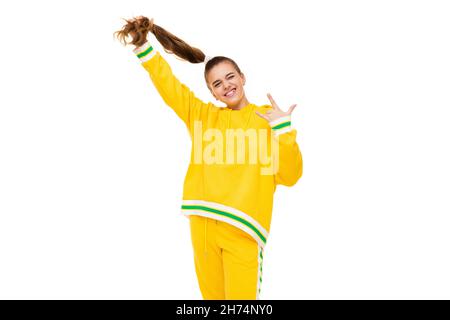 Photo d'une belle fille souriante avec des cheveux foncés avec une queue de cheval dans un élégant jaune tracksuit avec des rayures vertes montre la main rock isolée sur blanc Banque D'Images