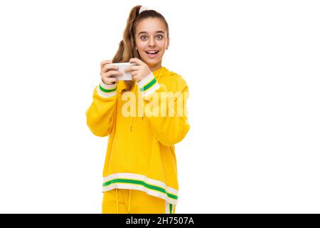 Photo d'une belle fille souriante avec des cheveux foncés avec une queue de cheval dans un élégant jaune tracksuit avec des rayures vertes joue un téléphone mobile avec un Banque D'Images