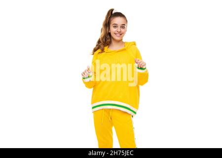 Beau drôle heureux merveilleux sourire mignonne fille avec des cheveux foncés avec queue de cheval dans élégant jaune tracksuit avec des rayures vertes isolées sur blanc Banque D'Images