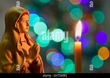 Composition de Noël : statuette de la Vierge Marie et une bougie d'église éclairée sur fond de lumières de Noël multicolores.Gros plan.Sélectionnez Banque D'Images