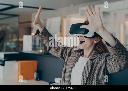 Impressionnée femme d'affaires aux cheveux rouges vêtue de lunettes VR portables explorant le monde virtuel Banque D'Images