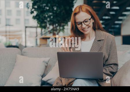 Heureuse femme d'affaires aux cheveux rouges agitant à la webcam d'ordinateur portable pendant la conférence Web avec le client Banque D'Images