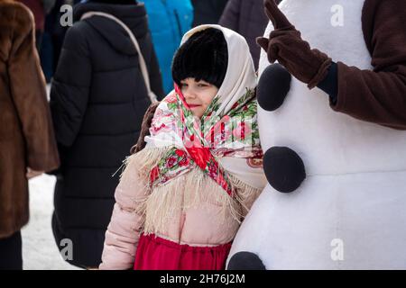 Une fille dans un foulard folklorique russe se tient près d'un acteur bonhomme de neige au festival adieu à l'hiver dans un parc de la ville. Banque D'Images