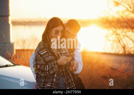 Le voyageur de couple se tient en voiture au coucher du soleil dans la campagne.Elle utilise la carte des applications mobiles dans son smartphone pour trouver le chemin.Les touristes qui vérifient la position dans la carte du smartphone.Mise au point douce Banque D'Images