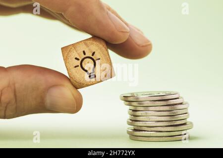 Un homme tient dans sa main un cube en bois avec une photo d'une ampoule près d'une pile de pièces de monnaie, un concept sur le sujet de l'économie d'énergie Banque D'Images
