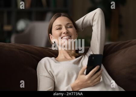Une femme se détendant sur un canapé tient le sourire de smartphone regarder l'appareil photo Banque D'Images