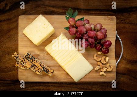 Tranche de fromage cheddar tranché sur une planche à bois avec des raisins, des noix de cajou, des bâtonnets de pain et du miel Banque D'Images